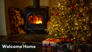 Omaze Million Pound House - Lake District House Draw. A fire burns inside a log burning stove with logs piled up next to it and a Christmas tree decorated with sparkly lights and baubles with presents beneath it